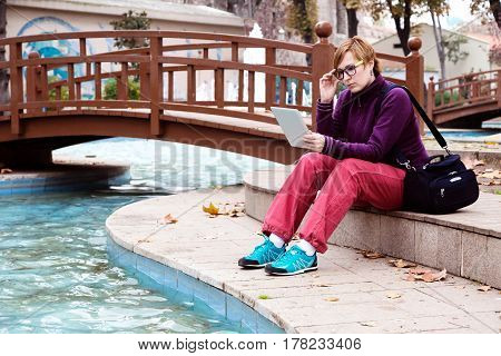 Cute Woman in casual and sporty Clothing sitting in Park next to Pond and using tablet pc device