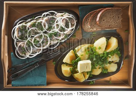 Pieces of a herring with rings of onions and fennel, hot boiled potatoes with green onions and a piece of butter on two black ceramic plates of an ovoidny form on a wooden tray from a bamboo