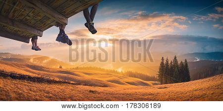 Hikers sit on a wooden flooring above the carpathian mountain valley and enjoy golden sunset.