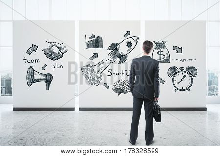 Back view of young businessman with briefcase looking at poster with business icons in interior with city view. Entrepreneurship concept. 3D Rendering