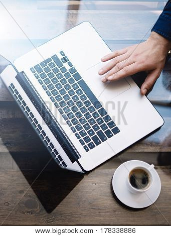 Man working with laptop at sunny office.Male hand pointing on notebook trackpad.Modern notebook, cup of black coffee on the wooden table.Reflections on glass surface.Vertical.Top view
