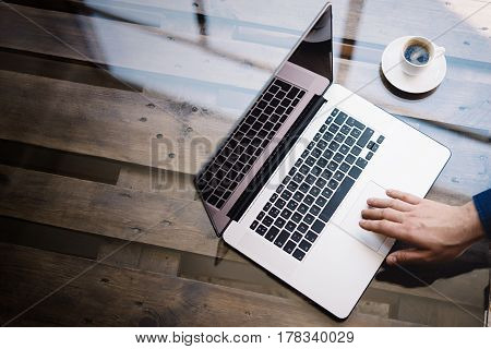 Man using laptop at sunny office.Male hand touching notebook trackpad.Modern notebook, cup of black coffee on the wooden table.Reflections on glass surface.Vertical.Top view