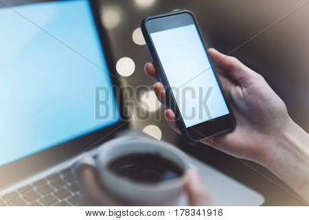 Hipster girl using computer and cup coffee in home atmosphere person holding blank smartphone on background glow illumination female hands texting on relax glitter bokeh mockup templates blur