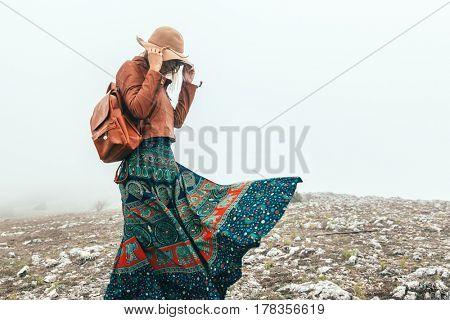 Boho woman wearing hat and leather jacket walking in mountains. Cold weather and fog. Spring or fall hiking. Wanderlust.