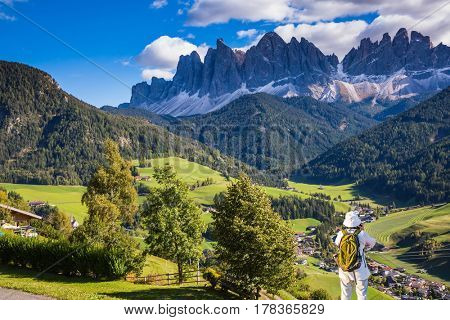 Woman-tourist with backpack photographs at Dolomites, Northern Italy. Forested mountains surrounded by green Alpine meadows and farm. The concept of active and eco-tourism