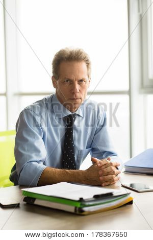 Angry businessman sitting at table and looking at camera in office. Boss of company going to have interview with new workers. Handsome man sitting with clasped hands.