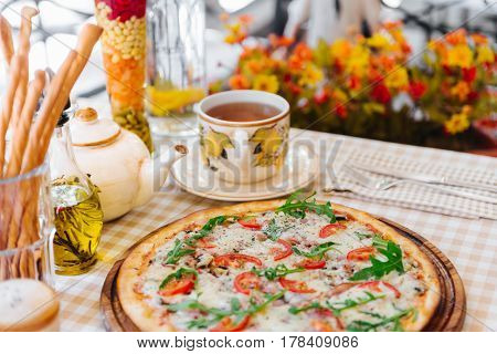 Pizza with cheese, tomato and meat, with cup of tee on served table with plaid tablecloth