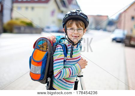 active school kid boy in safety helmet riding with his scooter in the city with backpack on sunny day. happy child in colorful clothes biking on way to school. safe way for kids outdoors to school.