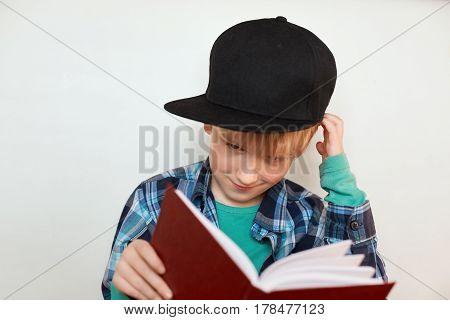 A portrait of liitle curious child with blond hair in stylish cap holding big red book in his hands scratching his head trying to understand what is written in the book. Children and education.