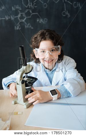 Exploring genome mutations. Happy capable brilliant child sitting in the laboratory and enjoying science lesson while makig notes and using microscope