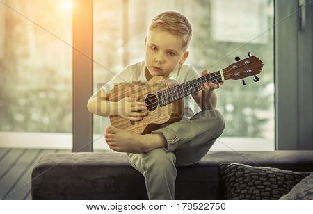 Young boy play on guitar at home at sunny day. Boy play on ukulele - hawaiian guitar.