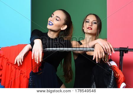 Two pretty girls or sexy women fashion models with long hair stylish makeup red and blue smiling lips in sexi black bodysuits shopping at clothes rack wardrobe on green or pink background