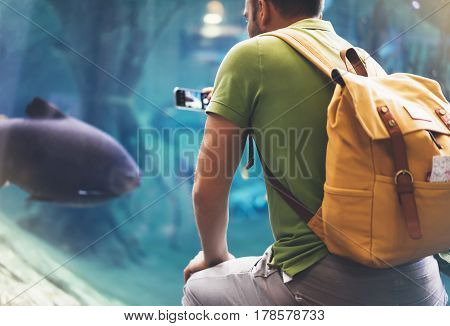 Hipster with yellow backpack map phone. View from back tourist traveler on background blue sea aquarium. Person hiker in oceanarium museum Barcelona and photographer pictures fish smartphone mockup