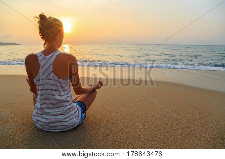 Young woman practices yoga and meditates in the lotus position on the beach at sunrise