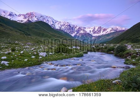From the river shore covered with stones opens view on fantastic glacier and steep rocky mountains with green meadows which are covered with snow.Happy lifestyle. Beautiful universe.