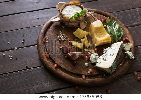 Cheese delikatessen on rustic wood. Blue cheese roquefort, parmesan, camembert and brie cuts decorated with garlic, pomegranate and basil, french cuisine ingredients