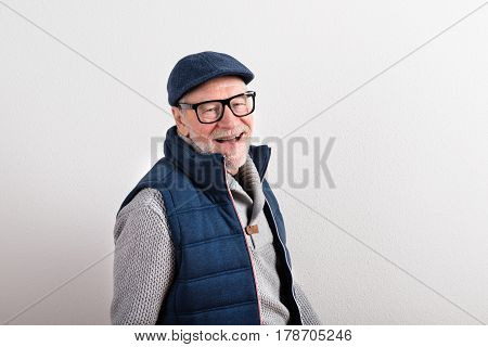 Handsome senior man in gray sweater, blue vest jacket, black eyeglasses and flat cap smiling. Studio shot against white wall.