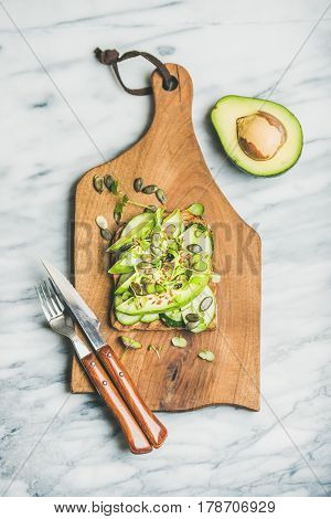 Healthy green veggie breakfast concept. Sandwich with avocado, cucumber, kale, kress sprout, pumpkin seeds on wooden board over marble background, top view. Vegan, detox, vegetarian, diet food concept