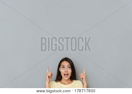 Image of surprised young woman dressed in yellow sweater posing over grey wall and looking aside to copyspace and pointing