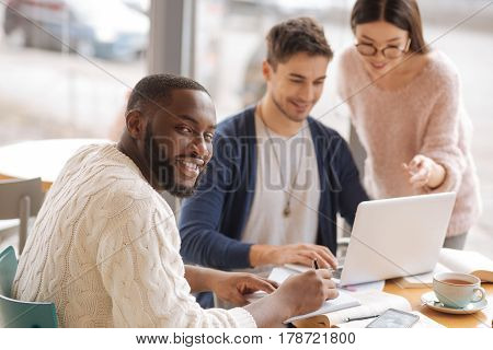 Happy startupper. Youthful bearded man is taking notes on background of his colleagues having discussion using laptop.