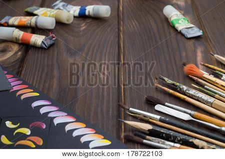 Art brushes and oil paints and sketches are laid out on a dark wooden surface