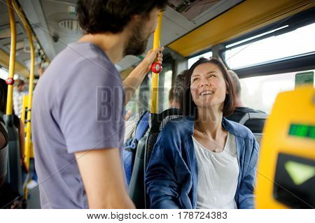 Young couple in inside of the city bus. The girl sits the fellow stands nearby. Young people look at each other and smile.