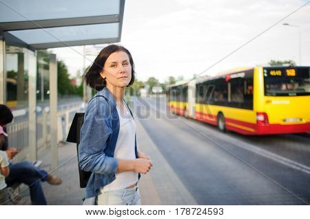 The young woman the brunette stands on the bus stop. She expects the bus. The woman is dressed in sports style. She high and slim. Behind shoulders at her a backpack.