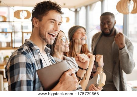 Extremely happy. Handsome youthful man is smiling and expressing happiness with his joyful fellows on background.