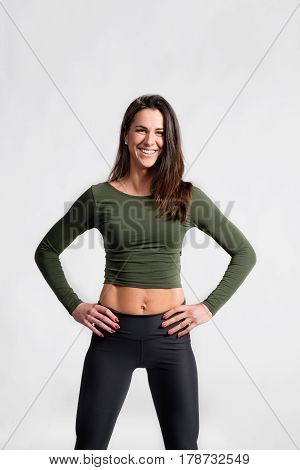 Attractive young fitness woman in green t-shirt and black sports leggings, arms on hips. Slim waist, perfect fit female body. Studio shot on gray background.