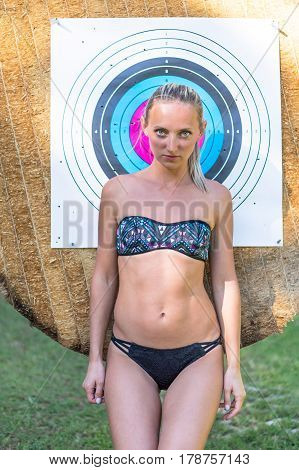 Attractive young smiling beautiful curly blonde hair slim girl fashion portrait in black swimming suit posing looking away calm look in target archery background.