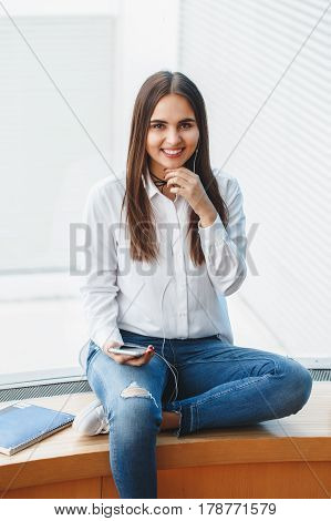 Portrait of smiling laughing young beautiful Caucasian girl woman in white shirt and blue jeans sitting on window sill in college university listening to music on phone