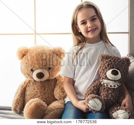 Charming little girl is looking at camera and smiling while sitting with her teddy bears at home