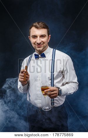 Portrait of a handsome smiling man in bow-tie and suspenders holding glass of beverage and cigar while looking at camera. Smoke on background. Studio shot