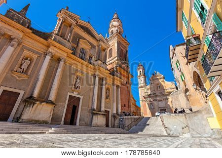 Small town square and Saint-Michel Archange Basilica under blue sky in Menton, France.