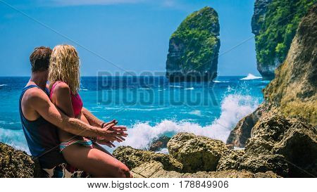 Couple in front of the Rock and watching Huge Waves hitting Tembeling Coastline at Nusa Penida Island, Bali Indonesia.