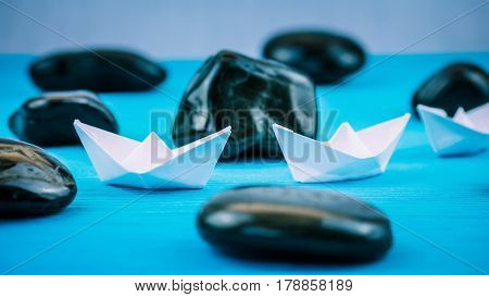Two White Paper Ships in Sigle File between Abstract Black Rock Stones on Blue Background. Side Shot.