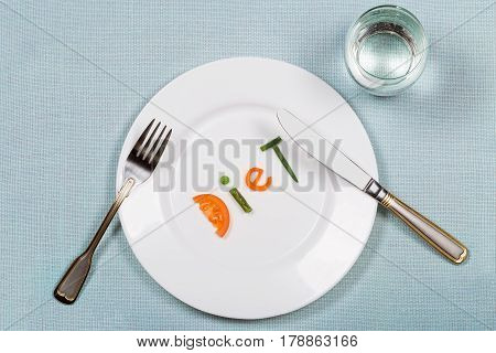 White plate with word diet made of colorful pieces of vegetables and glass of pure on blue teblecloth background. Flat lay. Top view. Concept of healthy eating and raw food dieting.