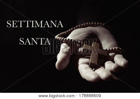 closeup of the hand of a young caucasian man with a rosary with a wooden cross in it, and the text settimana santa, holy week in italian, against a black background