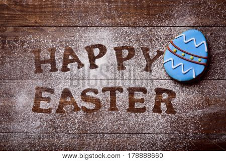 high-angle shot of a wooden table sprinkled with icing sugar or flour where you can read the text happy easter and a cookie decorated as an ornamented easter egg