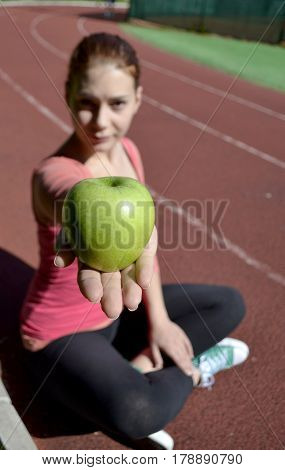 Young girl sitt on the running track and giving a green apple with her hand, hand and apple are focus