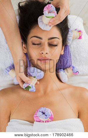 Herbal ball massage in ayurvedic spa. Female massagist with young woman in wellness center. Healthcare therapy to beautiful indian girl in beauty parlor, top view of face with eyes closed