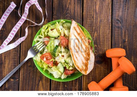 The chicken breast and salad on a table with dumbbells and measure tape. Top view studio shot. Copy space.