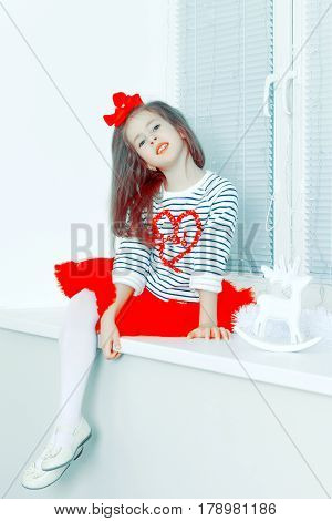 Beautiful little girl a bright red bow on head and red skirt sitting by the window.She put one leg over the other.Creative toning of a photograph.