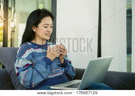 Happy young woman is relaxing on comfortable couch and using laptop at home. Photo toned.