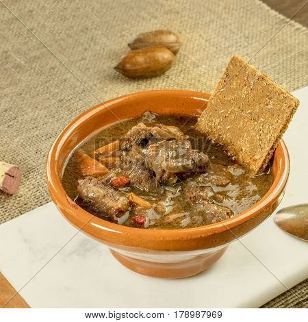 A square photo of a meat and vegetable stew in an earthenware bowl, with a flatbread and a wine cork
