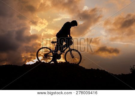 Silhouette Of A Boy Riding A Mountain Bike On The Hill At Yellow And Red Sunset Sky Color.