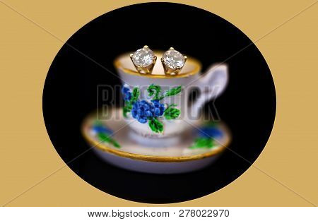 A Pair Of White Diamond Stud Earrings Set In Yellow Gold, Sitting In A Miniature Teacup On A Black O
