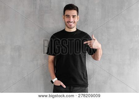 Young Handsome Man Isolated On Gray Textured Wall, Smiling While Pointing With Index Finger To Black