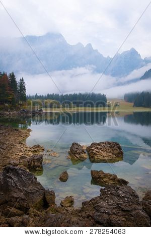 View Over Lake Laghi Di Fusine On A Foggy Morning With Some Rocks At The Beach And Mountain Range Ma