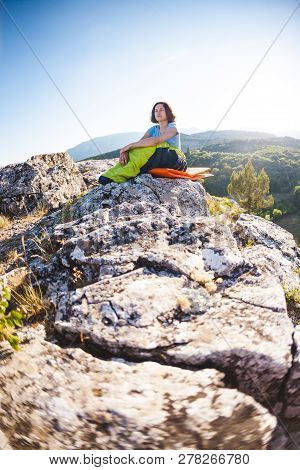 A Woman Is Sitting In A Sleeping Bag At Sunset. Camping On Top Of A Mountain. A Smiling Girl Rests A